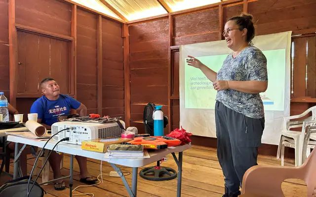 Irene Meulenberg teaching the community archaeology training in a wooden hall, beside a table of survey tools and a projected slide asking what tools an archaeologist needs