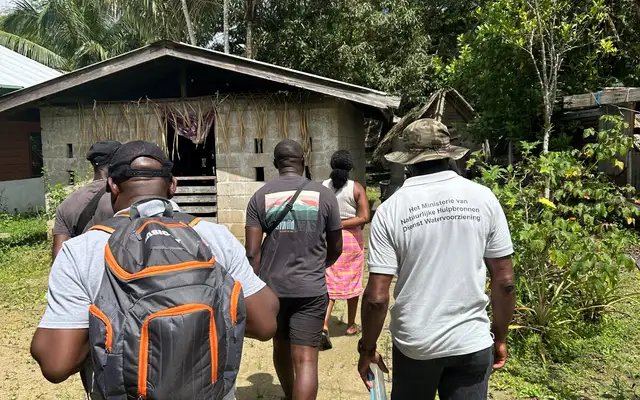 Members of the orientation visit walking through Tottikamp towards a breeze-block house, one of them in a shirt of the Ministry of Natural Resources' Dienst Watervoorziening